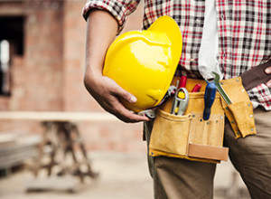 Gros plan sur un homme ayant un ceinture d'outils de construction et son casque dans la main, accoté sur sa hanche.
