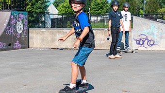 Jeune garçon portant un casque et se déplaçant en planche à roulettes dans un roulodrome. Deux garçons le regarde au loin.
