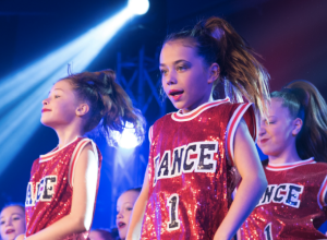 Trois jeunes filles sur scène à l'occasion d'un spectacle de danse, toutes vêtues d'une camisole sportive à paillettes rouges portant l'inscription DANCE.