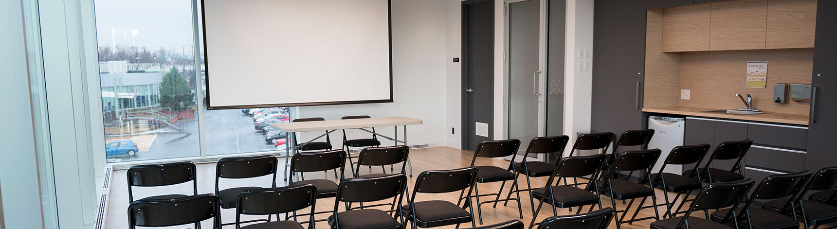 Salle vitrée avec un écran pour projecteur, 3 rangées de chaises noires pliantes et espace de service avec comptoir et lavabo.